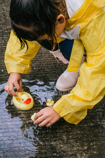 Encantador niño étnico en impermeable jugando con patos de plástico que se reflejan en charco ondulado en tiempo lluvioso - foto de stock
