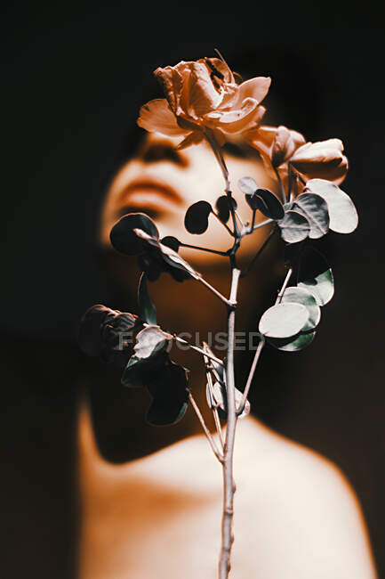 Mujer joven tierna con sombra en la cara contra la flor en flor en tallo delgado con follaje sobre fondo negro - foto de stock