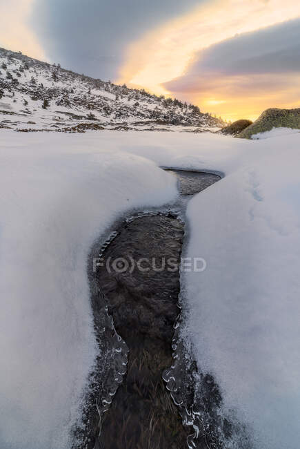 Paisagem espetacular de rio frio fluindo entre terreno nevado em área montanhosa sob céu nublado colorido ao nascer do sol — Fotografia de Stock