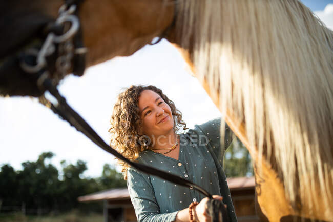 Content mature female stroking stallion muzzle with smooth brown coat against hay in riding school on sunny day — Stock Photo