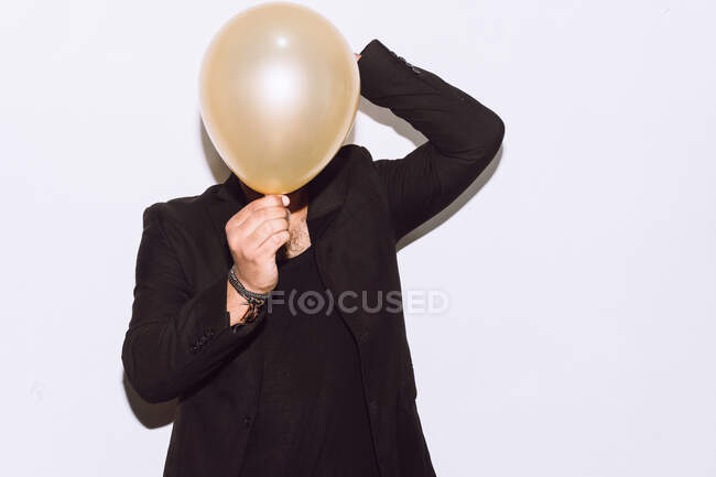 Anonymous male in black outfit hiding face behind balloon during holiday celebration against white background — Stock Photo