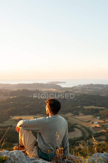 Вид ззаду на невпізнаваного мандрівника-чоловіка, який захоплюється океаном і плантаціями з деревами, відпочиваючи на хребті під час заходу сонця — стокове фото