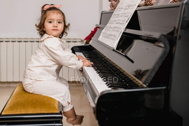 Vista lateral del encantador niño descalzo en pijama mirando hacia adelante mientras toca el piano en casa - foto de stock