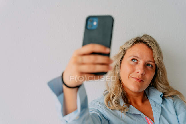 Mulher loira adulta com cabelo ondulado tirando selfie no celular enquanto tem tempo livre no fundo claro — Fotografia de Stock