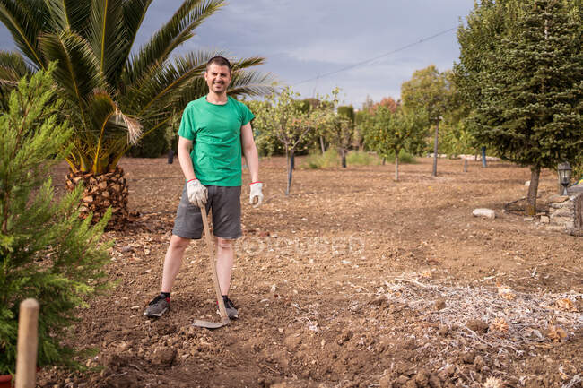 Adult male horticulturist standing looking at camera with hoe preparing soil to plant pine tree against mountains in daylight — Stock Photo