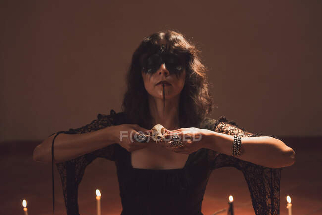 Dreamy enchantress with painted face and skull casting spell during mystic ritual in room with dim light — Stock Photo