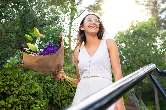 De baixo conteúdo jovem fêmea em óculos olhando para longe de pé com buquê de flores florescentes em escadas urbanas — Fotografia de Stock