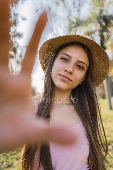 Усміхнена жінка-підліток з брекетами, що демонструють фотографічний жест, дивлячись на камеру вдень на розмитому фоні — стокове фото