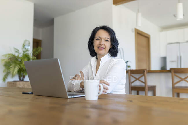 Feliz madura freelancer mujer navegando en Internet en netbook trabajando en un nuevo proyecto mientras está sentado en la mesa en casa - foto de stock
