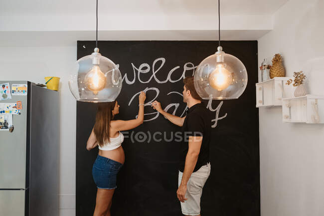 Male writing Welcome Baby inscription on blackboard against expectant female beloved in house — Stock Photo