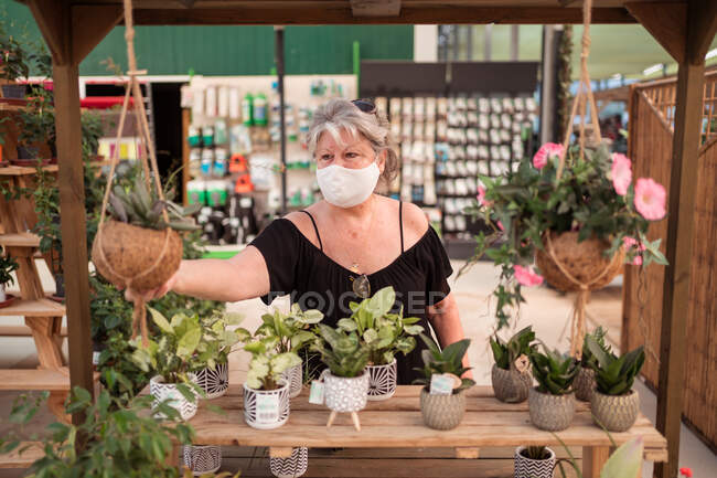 Зрелая женщина-покупатель в текстильной маске собирает растения в горшках во время пандемии коронавируса в садовом магазине — стоковое фото
