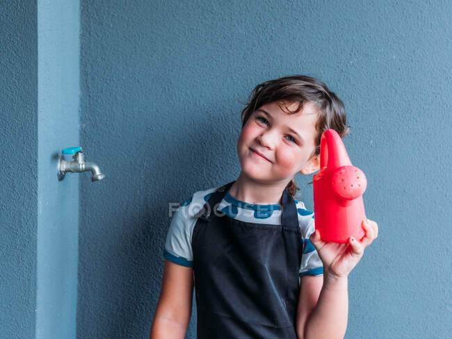 Happy little girl standing near gray wall and showing the watering can in daytime while looking at camera — Stock Photo