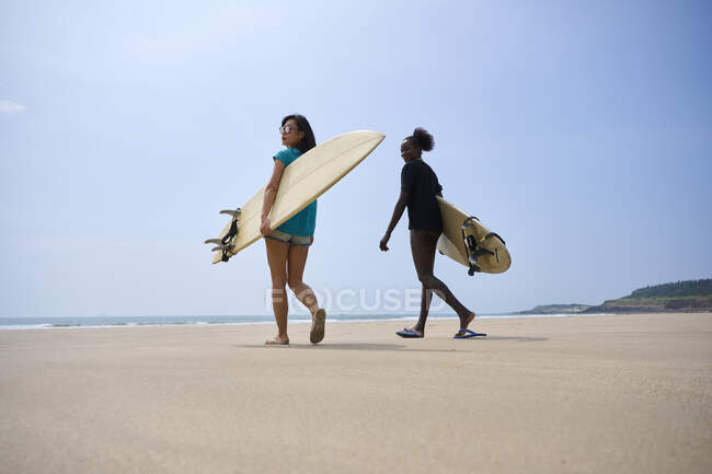 Vista posterior de jóvenes surfistas femeninas multiétnicas con tablas de surf hablando mientras pasean por la orilla arenosa y miran hacia otro lado - foto de stock