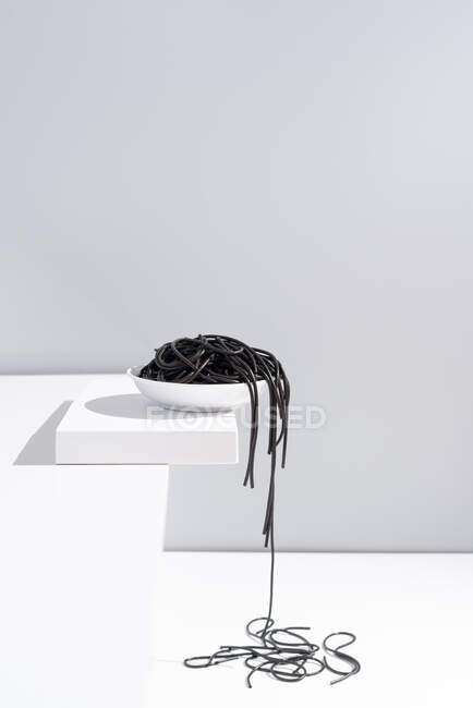 Estúdio minimalista com espaguete de tinta de lula preta caindo da tigela de cerâmica completa na mesa branca — Fotografia de Stock