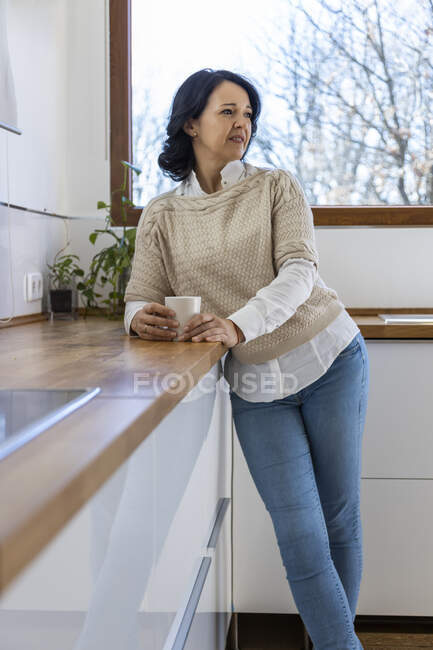 Зрелая уверенная женщина, стоящая в стороне на кухне возле столешницы держа кружку горячего напитка утром дома — стоковое фото