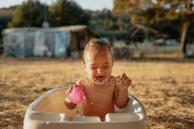 Criança feliz com olhos fechados e brinquedo sentado em banho de plástico enquanto brincava com a água no campo — Fotografia de Stock