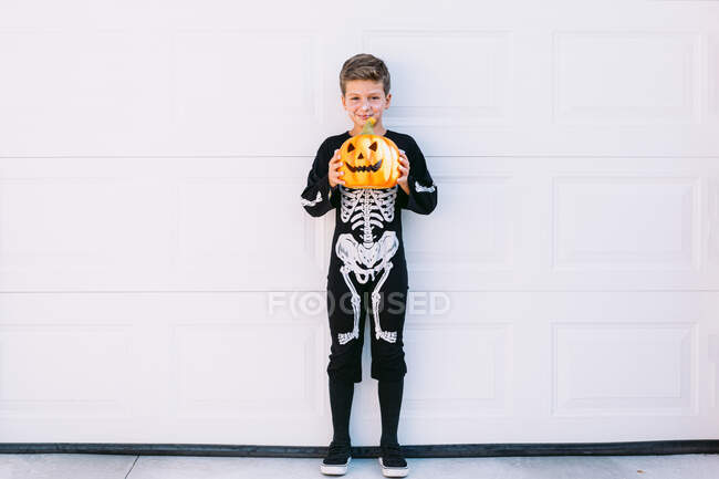 Corpo inteiro de menino pré-adolescente sorridente vestindo traje de Halloween preto com impressão de esqueleto em pé perto esculpida Jack O Lanterna abóbora contra a parede branca — Fotografia de Stock