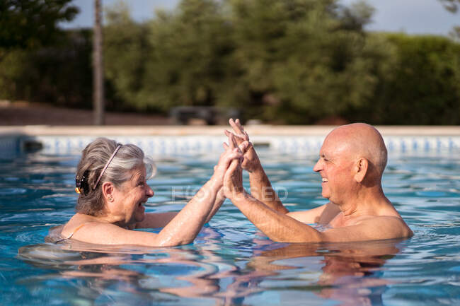 Side view of cheerful senior couple holding hands while swimming in pool and looking at each other — Stock Photo