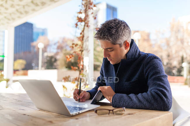 Side view of concentrated young bearded male freelancer in casual clothes and eyeglasses sitting at table with laptop and smartphone while working on project in outdoor cafe — Stock Photo
