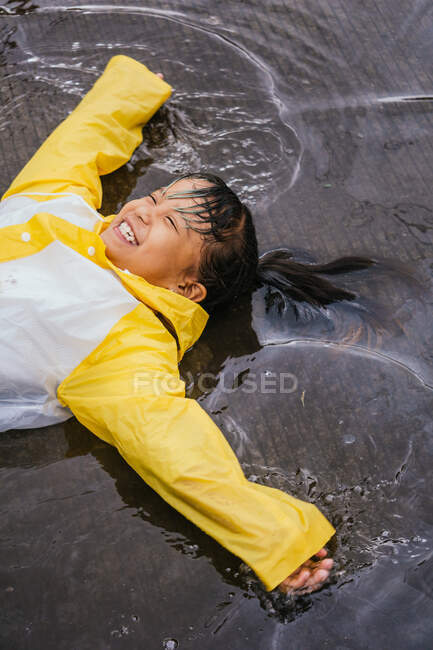 Asiatisches Kind in Gummistiefeln und Slicker liegt an Regentagen in Pfütze — Stockfoto