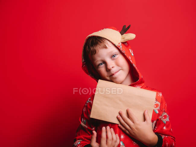 Glücklicher kleiner Junge im festlichen Kostüm, der bei der Weihnachtsfeier vor rotem Hintergrund in die Kamera schaut — Stockfoto