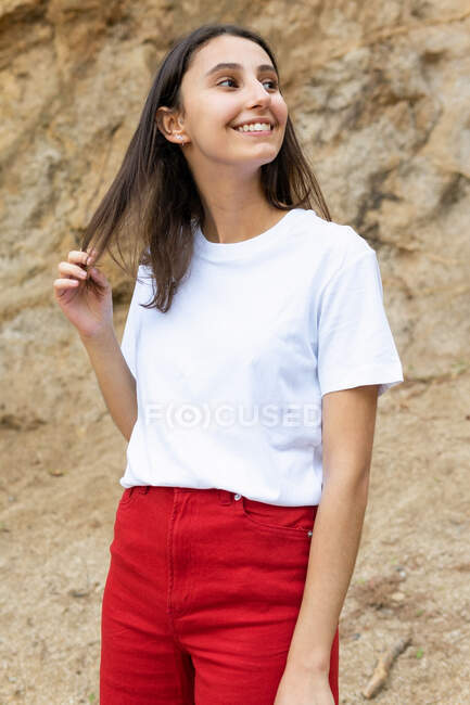 Young contemplative happy female adolescent in white t shirt and red jeans looking away while standing on rough land against mount — Stock Photo