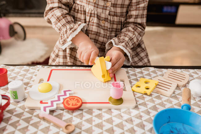Crop unrecognizable kid in checkered dress with toy knife cutting cheese on chopping board while playing in house — Stock Photo