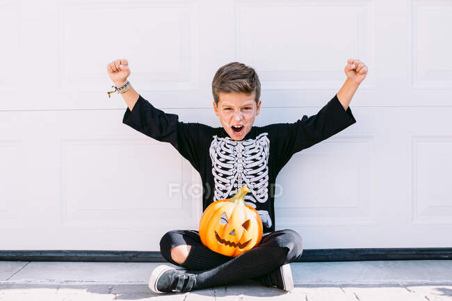 Full body of excited boy in skeleton costume with makeup and carved Halloween pumpkin raising arms and screaming with scary face while sitting near white wall — Stock Photo