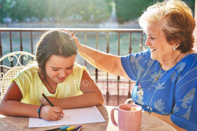 Lächelnde Großmutter streicht über den Kopf der fröhlichen Enkelin, die auf Papier zeichnet, während sie bei sonnigem Wetter die Zeit zusammen auf der Terrasse verbringt — Stockfoto