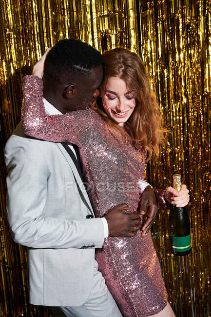 Amable joven con botella de champán abrazando a un hombre afroamericano amado mientras él la abraza y le dice cosas al oído durante la fiesta - foto de stock