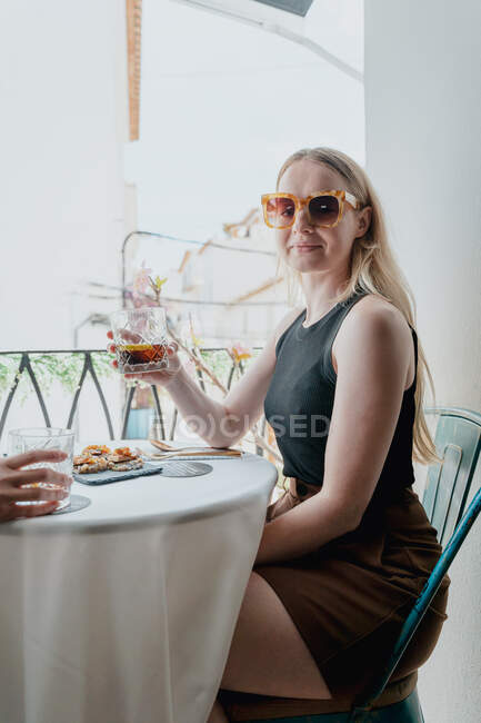 Вид сбоку на молодую женщину в солнечных очках, сидящую со стаканом виски за столом с другом урожая на балконе — стоковое фото