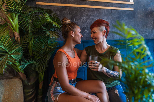 Contenido joven mujer tatuada con mohawk y bebida abrazando novia lesbiana mientras se miran en el sofá en casa - foto de stock