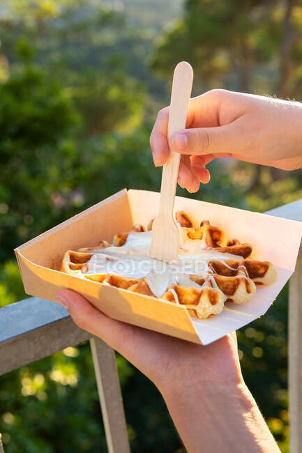 Ritagliato persona irriconoscibile mangiare gustosi waffle belgi con panna montata in scatola da asporto contro supporti in retroilluminato — Foto stock