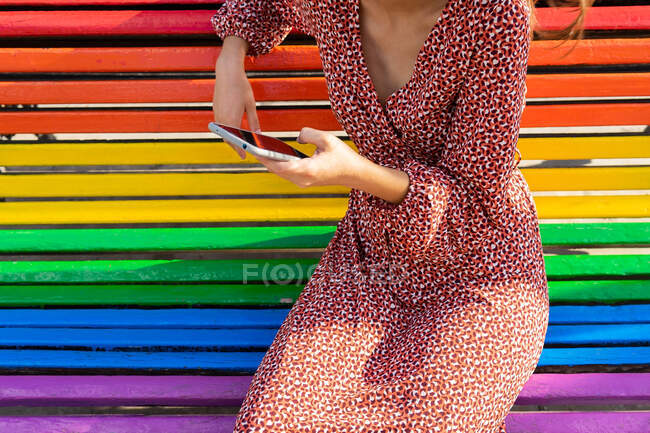 Cultiver femme méconnaissable en robe assis sur banc coloré et navigation téléphone mobile dans la journée — Photo de stock