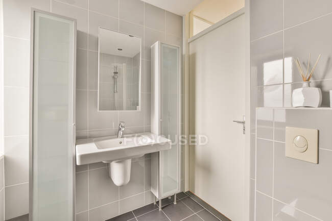 Armarios de baño y lavabo de cerámica bajo el espejo contra el cuarto de baño y palos aromáticos en jarrón en el estante en casa - foto de stock
