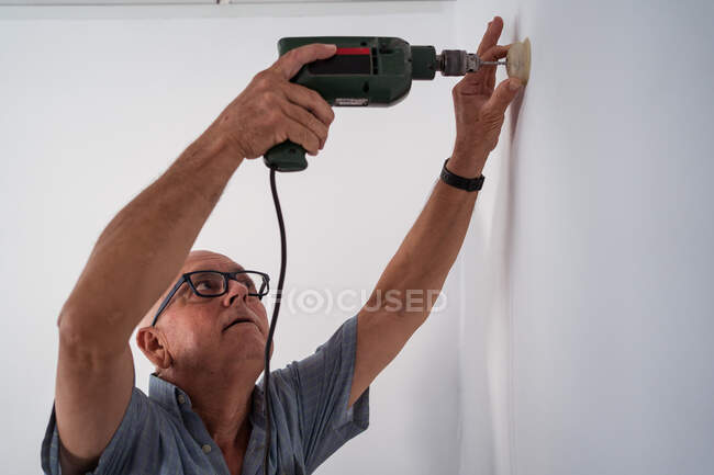 Concentrato vecchio maschio in occhiali con cacciavite elettrico avvitando pezzo di plastica a parete in casa — Foto stock