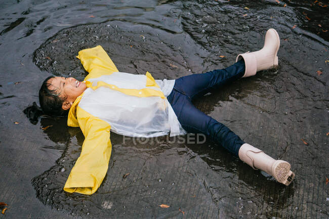 Asiatisches Kind in Gummistiefeln und Slicker liegt an Regentagen in Pfütze — Stockfoto