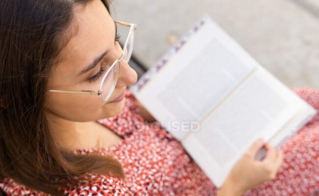 Зверху позитивна молода жінка в стильному одязі сидить з відкритою книгою на дерев'яній лавці проти будівлі зі світлою стіною вдень — стокове фото