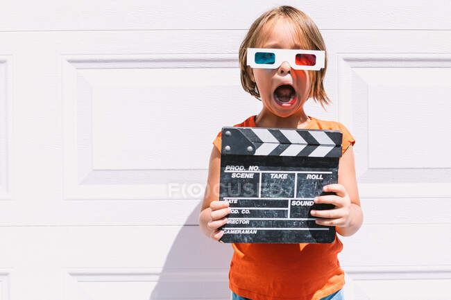 Cute astonished girl in casual colorful clothes holding clapperboard while on three dimensional glasses standing on white wall background — Stock Photo