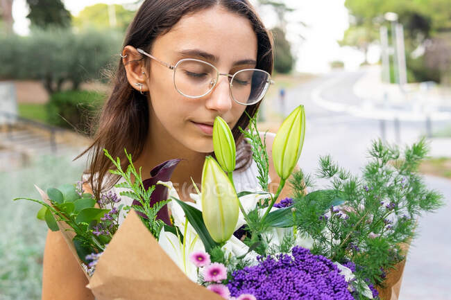 Заботливая молодая женщина с закрытыми глазами в очках наслаждается ароматом цветущего цветочного букета в городе на размытом фоне — стоковое фото