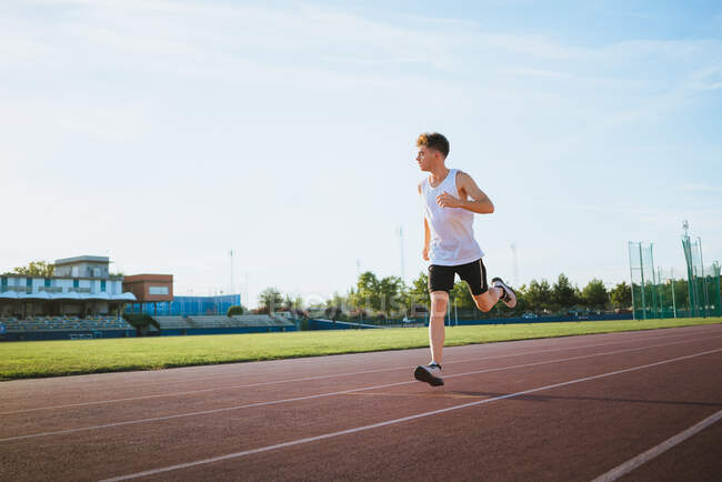 Deportista activo corriendo mientras mira hacia otro lado durante el entrenamiento en pista bajo el cielo nublado en la ciudad soleada - foto de stock