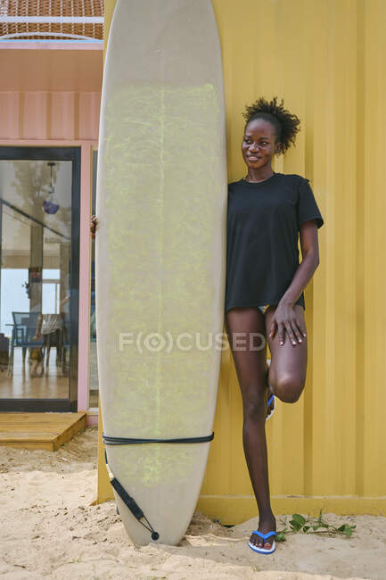 Joven deportista afroamericana alegre en bikini y camiseta mirando hacia otro lado con tabla de surf en un bar de playa en la costa - foto de stock