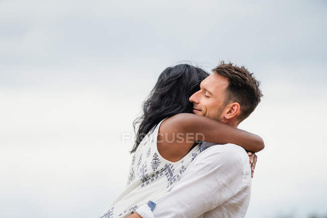 Vista lateral del hombre sonriente abrazando novia india de pie en el campo bajo el cielo nublado - foto de stock