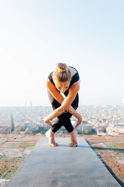 Young barefoot female in sportswear leaning forward with crossed legs while practicing yoga on rooftop in sunlight — Stock Photo