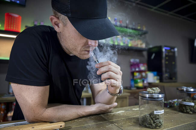 Anonymous adult male in cap smoking cannabis joint in workspace on blurred background — Stock Photo