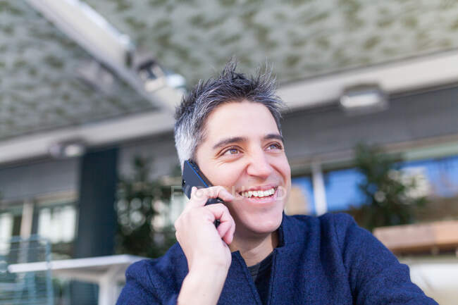 Счастливый молодой человек в повседневной одежде улыбается, сидя на скамейке и отвечая на телефонный звонок — стоковое фото