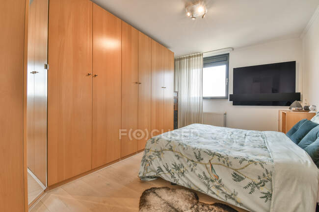 Creative design of bedroom with bed against wooden closet and television above parquet with carpet in house — Stock Photo