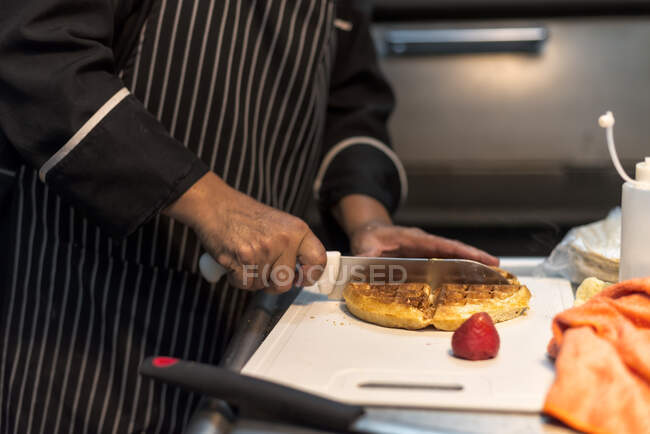 Невідомий чоловічий кухар з вирізанням ножа Бельгійський вафель на дошці рубають з свіжою полуницею на роботі. — стокове фото