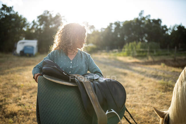 Femme avec selle et cheveux bouclés contre l'étalon mangeant de l'herbe à la campagne dans le dos éclairé — Photo de stock