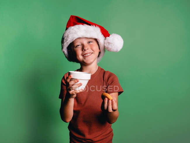Чарівний маленький хлопчик з різдвяним капелюхом Санта, беручи печиво з чашки на зеленому тлі, дивлячись на камеру — стокове фото
